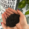 Hand holding a portion of activated biochar with a package labeled 'Activated & Inoculated Biochar' in the background.
